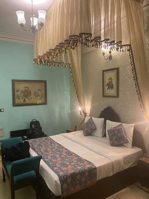 A richly decorated hotel bedroom with pastel walls, framed artwork, a canopy, and patterned pillows on a double bed.