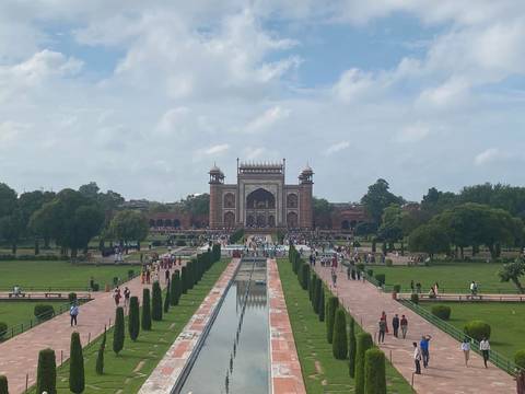 A broad view of the grand gateway to the Taj Mahal complex with long reflecting pools and manicured gardens bustling with visitors.