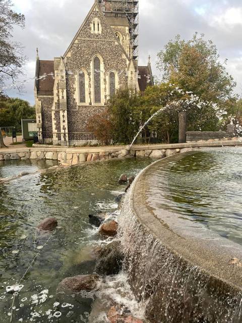 A graceful fountain arches water into a stone basin near a historic building and greenery.