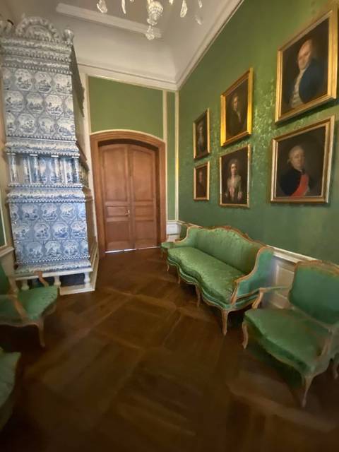 A slightly blurry palace room with green damask walls, antique portraits, and ornate tiled stove.