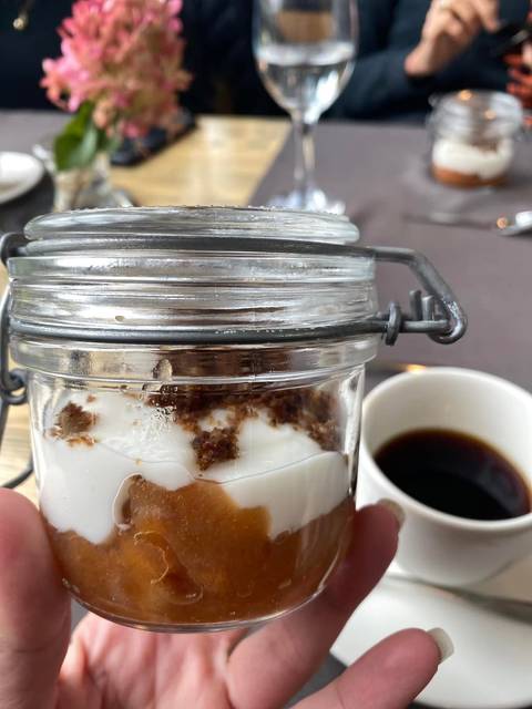 A close-up of layered dessert in a glass jar beside a cup of black coffee on a table.