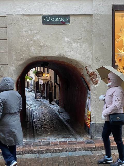 Cobblestone alley through an archway with pedestrians in raincoats and umbrellas.