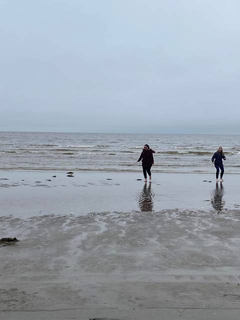 Two young women walk barefoot in the shallow water on a grey deserted beach.