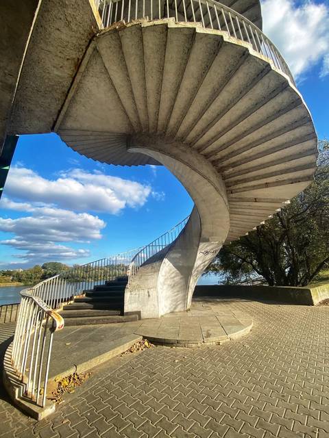 Artistic concrete spiral staircase curving above a river promenade under a blue sky.