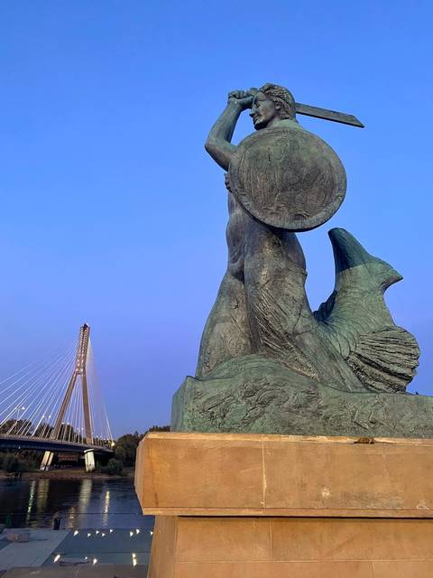 Iconic bronze Warsaw mermaid statue with cable-stayed bridge in background against clear sky.