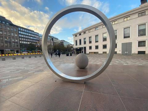 Modern outdoor sculpture of a metal ring framing an egg-shaped stone in a city plaza.