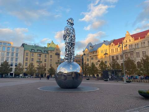 Reflective sphere sculpture topped by a perforated human figure in a European square at golden hour.