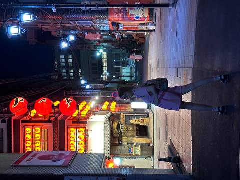 Young woman poses on an illuminated street lined with red lantern signs in a Japanese nightlife district.