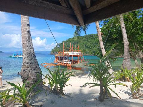 Traditional outrigger tour boat anchored at white-sand cove fringed by palms and turquoise water.