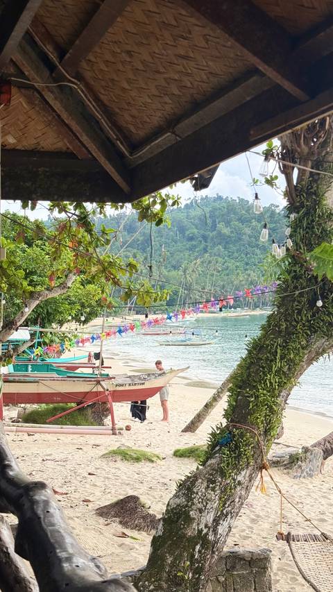 Coastal village scene with boats moored by beach, colourful paper pinwheels strung across lush greenery.