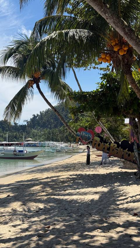 Beachfront walkway lined with boats and palm trees featuring a colourful 'Port Barton' heart sign.