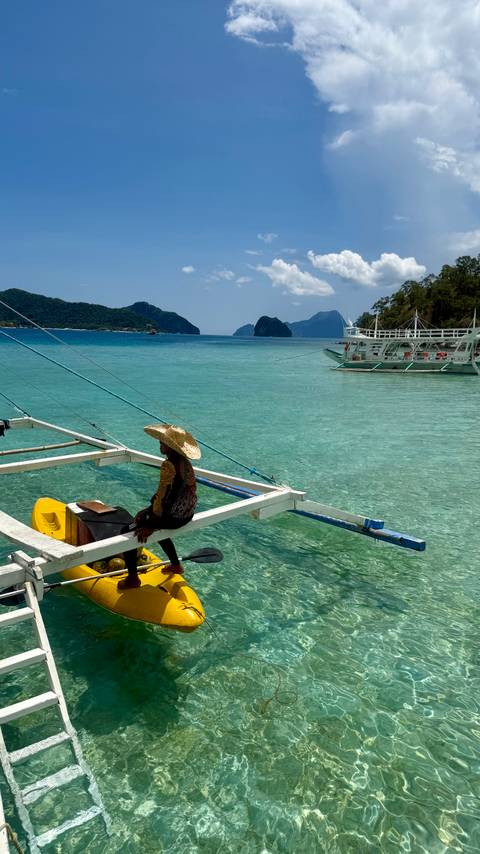 Tattooed man wearing a straw hat sits on the outrigger frame of a white banca boat looking over turquoise sea with dramatic limestone islands in the distance.