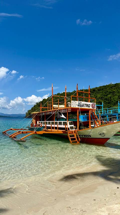 Bright wooden outrigger tour boat moored in clear blue water with green forested hills and blue sky.