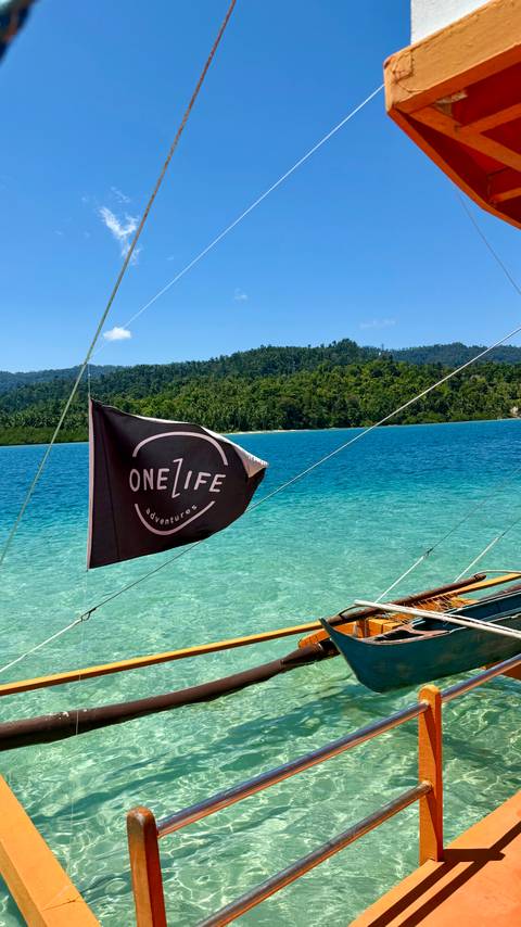Black flag reading 'One Life adventures' flutters from boat rigging above vibrant turquoise water and jungle-covered shoreline.
