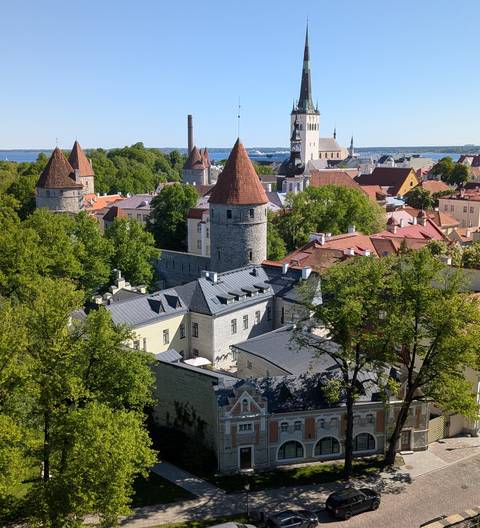 Scenic elevated view over Tallinn Old Town with stone towers, red-tile rooftops and the Baltic Sea in the background under clear blue skies.