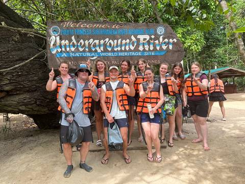 Group of smiling travellers wearing orange life vests posing in front of the Puerto Princesa Underground River entrance sign.