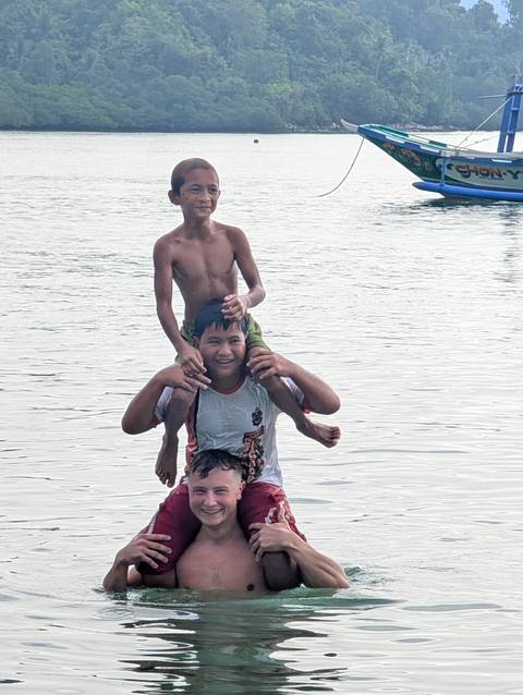 Three playful boys enjoying the water, one sitting on another’s shoulders against a calm sea backdrop.