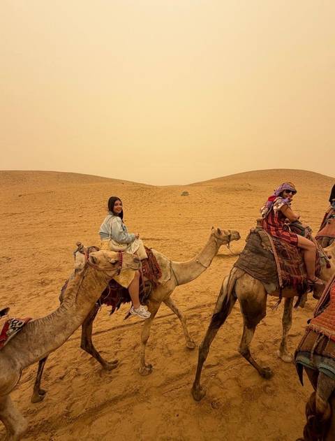 Travellers riding camels across golden desert dunes under a hazy sky near the pyramids.