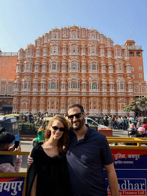 Couple standing before the ornate pink façade of Jaipur’s Hawa Mahal amid bustling street life.