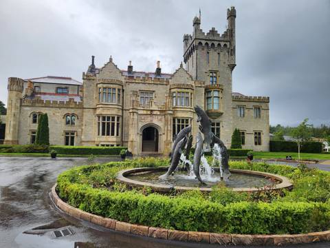 Historic castle hotel with dolphin fountain surrounded by manicured gardens on a rainy day.