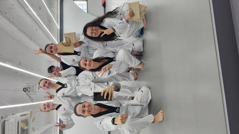 Seven participants in taekwondo uniforms pose indoors, holding broken boards and making victory signs.