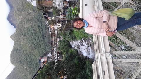 Traveler smiles on a bridge above a flowing mountain stream with town buildings and lush hills behind.