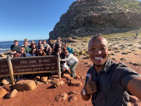 Large tour group posing for a selfie beside the Cape of Good Hope sign on a sunny coast.