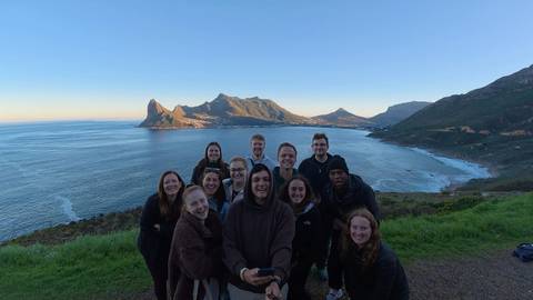 Smiling travelers take a selfie on a grassy viewpoint overlooking a wide bay and rugged mountains.