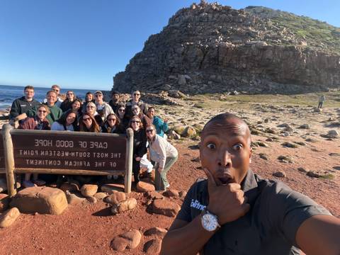 Energetic group gathered by the Cape of Good Hope sign while a guide pulls a playful face in the foreground.
