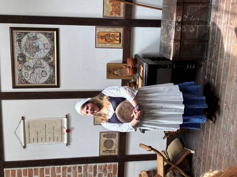 Costumed interpreter in medieval dress holds a loaf in a historic interior lined with maps and artifacts.