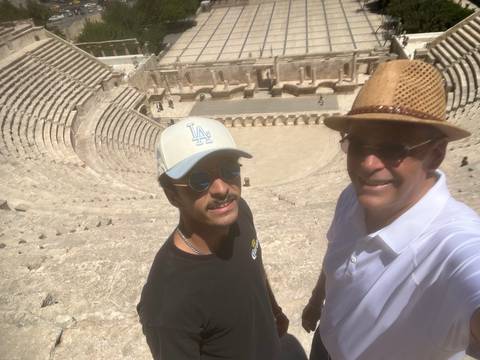 Father and son take a selfie high in the stone tiers of a large ancient Roman amphitheatre under bright sun.