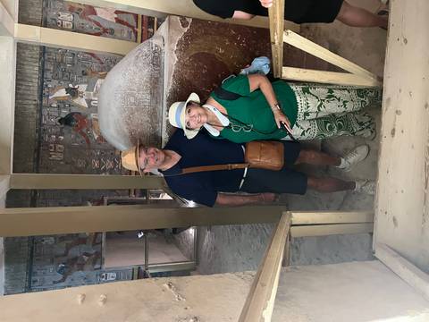 Smiling couple pose inside an ancient Egyptian tomb beside a massive stone sarcophagus decorated with art.