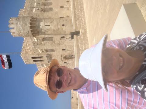 Smiling couple take a selfie in front of a stone coastal fortress flying the Egyptian flag.