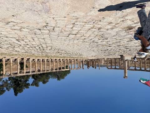Expansive oval forum of Jerash framed by standing Roman columns with a Jordanian flag fluttering and visitors exploring.