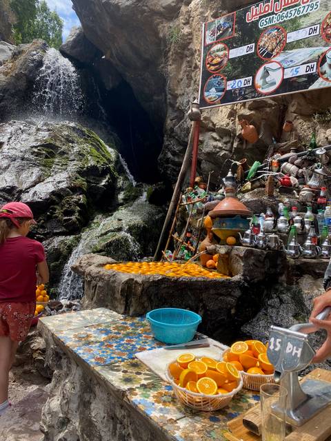 Small refreshment stall with oranges beside a cascading waterfall in the mountains.