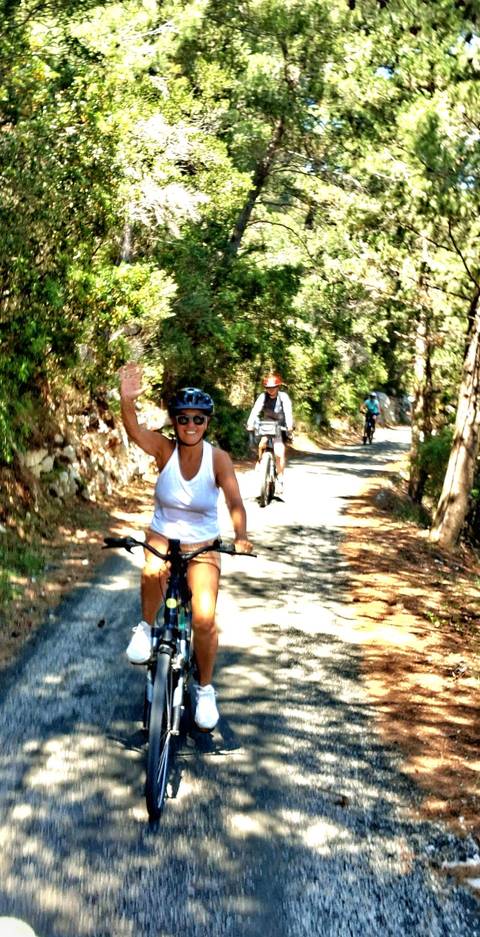Cyclists ride through a shady forest path as one rider waves to the camera.