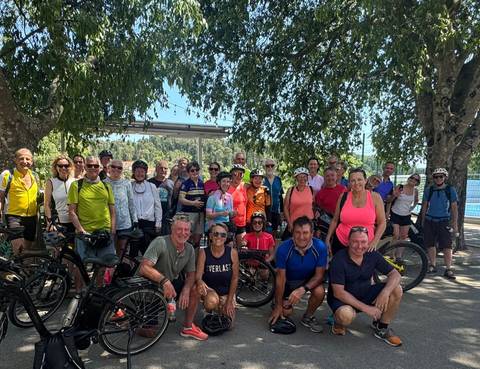 Large cycling tour group poses together with their bikes under leafy trees.