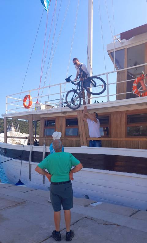 Crew hoists a bicycle onto a wooden boat while passengers watch.