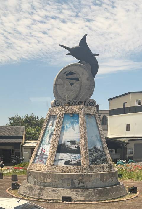 A sculptural monument topped with a round stone artwork against a blue sky; photos are displayed on its tiled base.