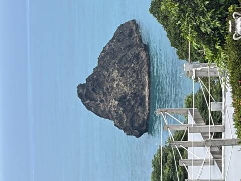 A rugged sea stack rises from calm turquoise waters near a lush coastline.