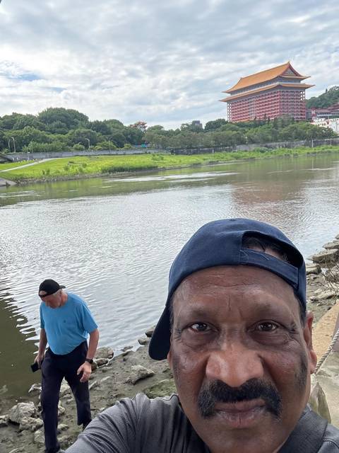 Partial selfie showing the top of a man’s head by a riverbank while another person looks at the water behind him.