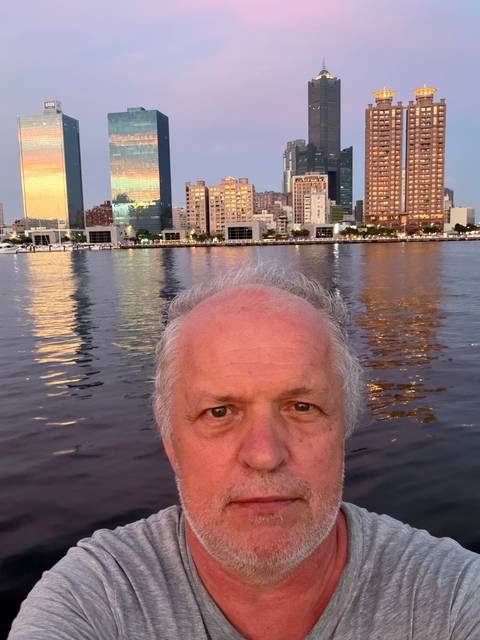 Close-up selfie of a man with a lit city waterfront skyline reflecting on calm water at dusk.