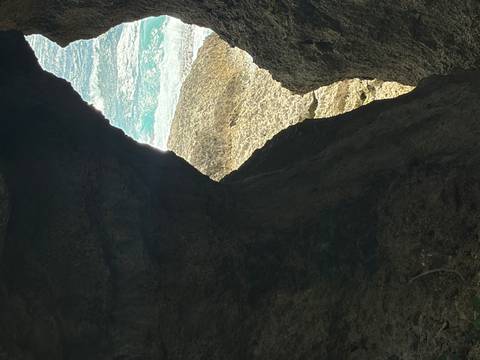 View through a dark rocky cave opening toward bright turquoise waves crashing on rocks.