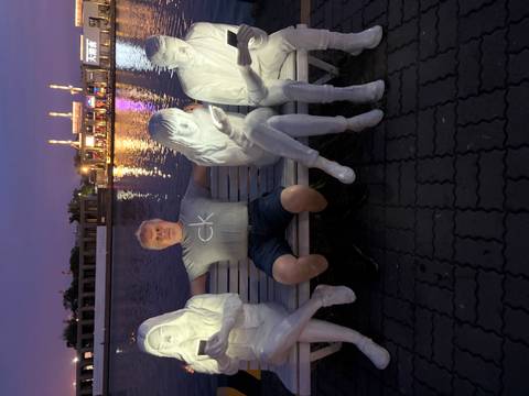 A man poses on a bench flanked by illuminated white statues beside calm evening water.