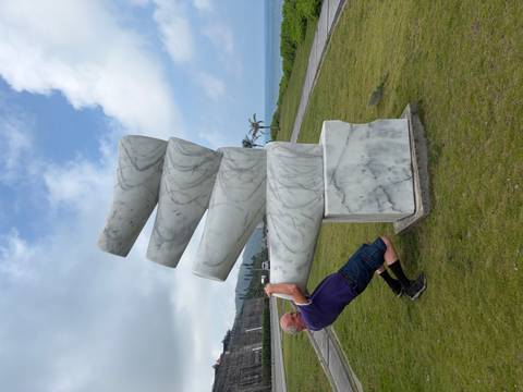 A man playfully pretends to push over a stack of leaning marble cylinders by the seaside.