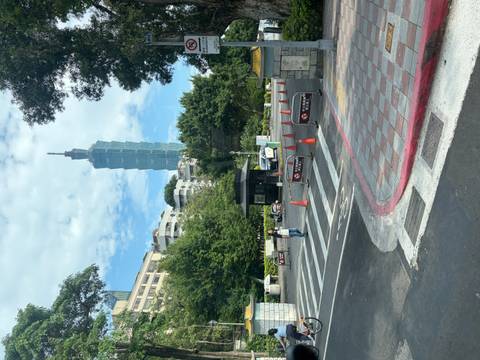 Street scene with Taipei 101 rising above trees and buildings beyond a crosswalk and traffic cones.
