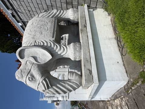 Grey stone elephant sculpture with decorative carvings outside a temple complex under blue sky.