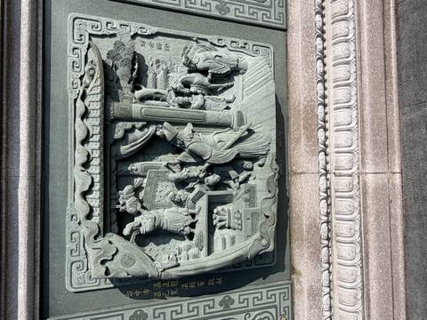 Sun-lit relief carving on green stone portraying an intricate domestic scene with figures and furnishings.