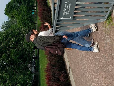 A young man and woman walk together along a paved park path beside a metal railing and greenery.