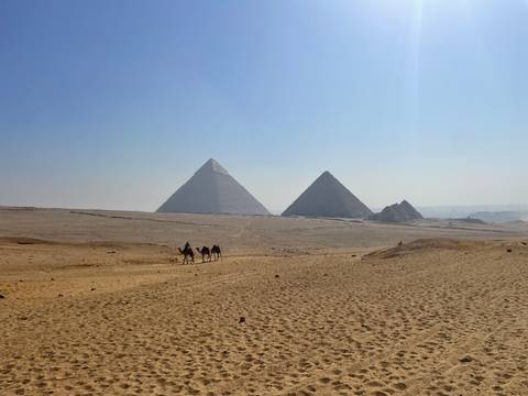 Wide desert vista with two main pyramids on the horizon and a small caravan of camels crossing the sands.